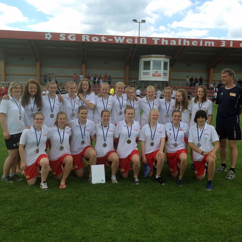 Eine Gruppenfoto von weiblichen Fußballspielern in weißen T-Shirts und roten Shorts, die Medaillen tragen und auf einem Sportfeld posieren.