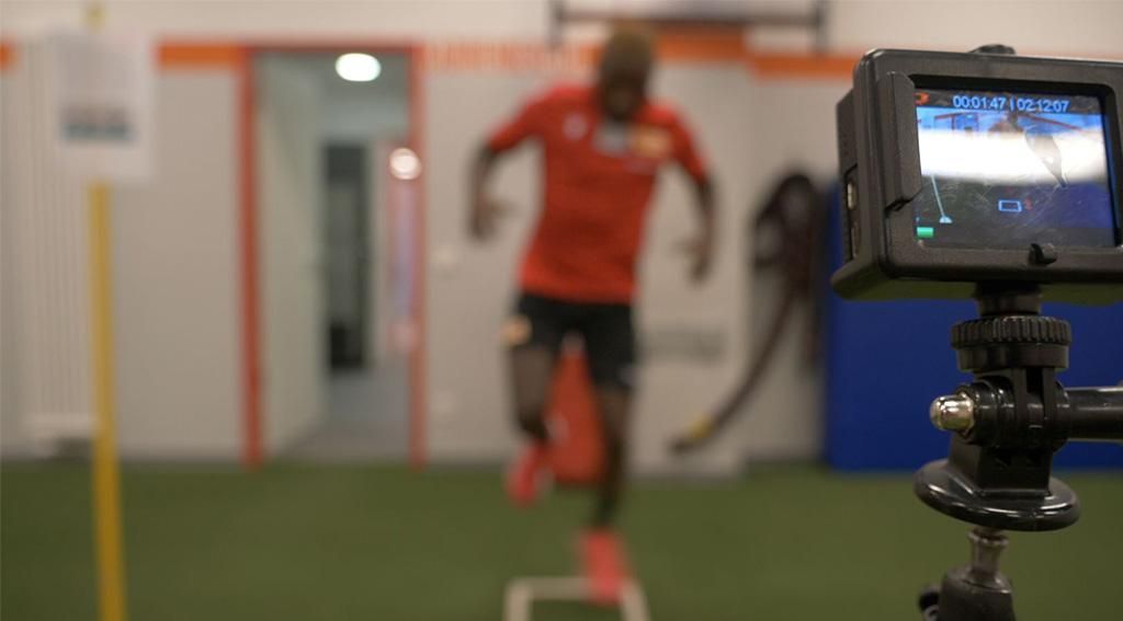 Ein Sportler in rotem Trikot springt über eine Trainingsleiter, während eine Kamera seine Leistung aufzeichnet.