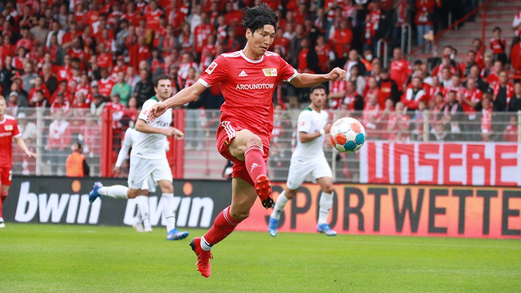 Fußballspieler in rotem Trikot schießt den Ball, während Fans im Hintergrund jubeln.