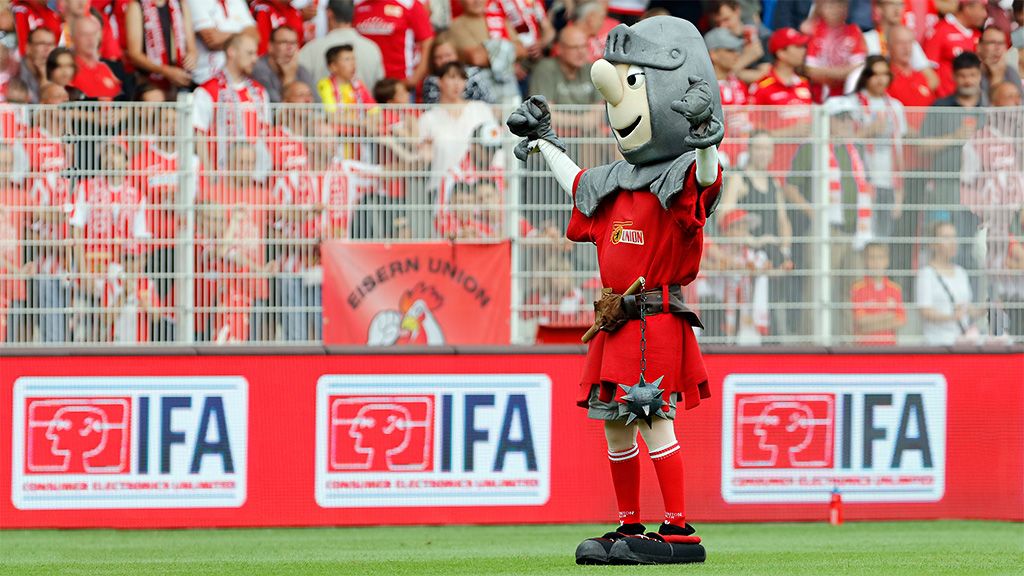 Mascot in knight costume with raised arms in front of cheering crowd in stadium wearing red and white fan gear.