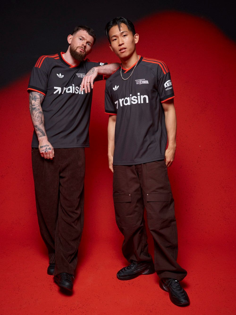 Zwei Männer stehen in sportlichen Outfits mit schwarzen Trikots vor einem roten Hintergrund.