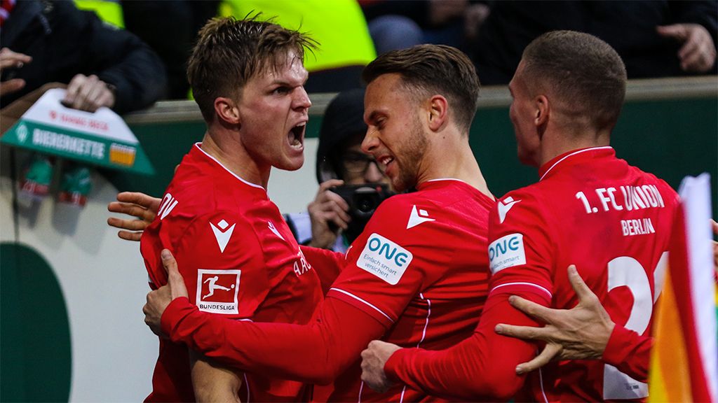 Drei Fußballspieler in roten Trikots freuen sich. Sie umarmen sich. Im Hintergrund feiern die Fans.