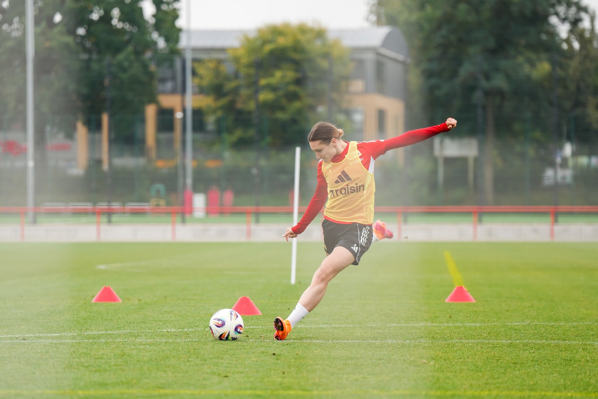 Eine Spielerin in roten und gelben Trainingsbekleidung schießt einen Fußball auf einem Trainingsplatz, umgeben von Hütchen.