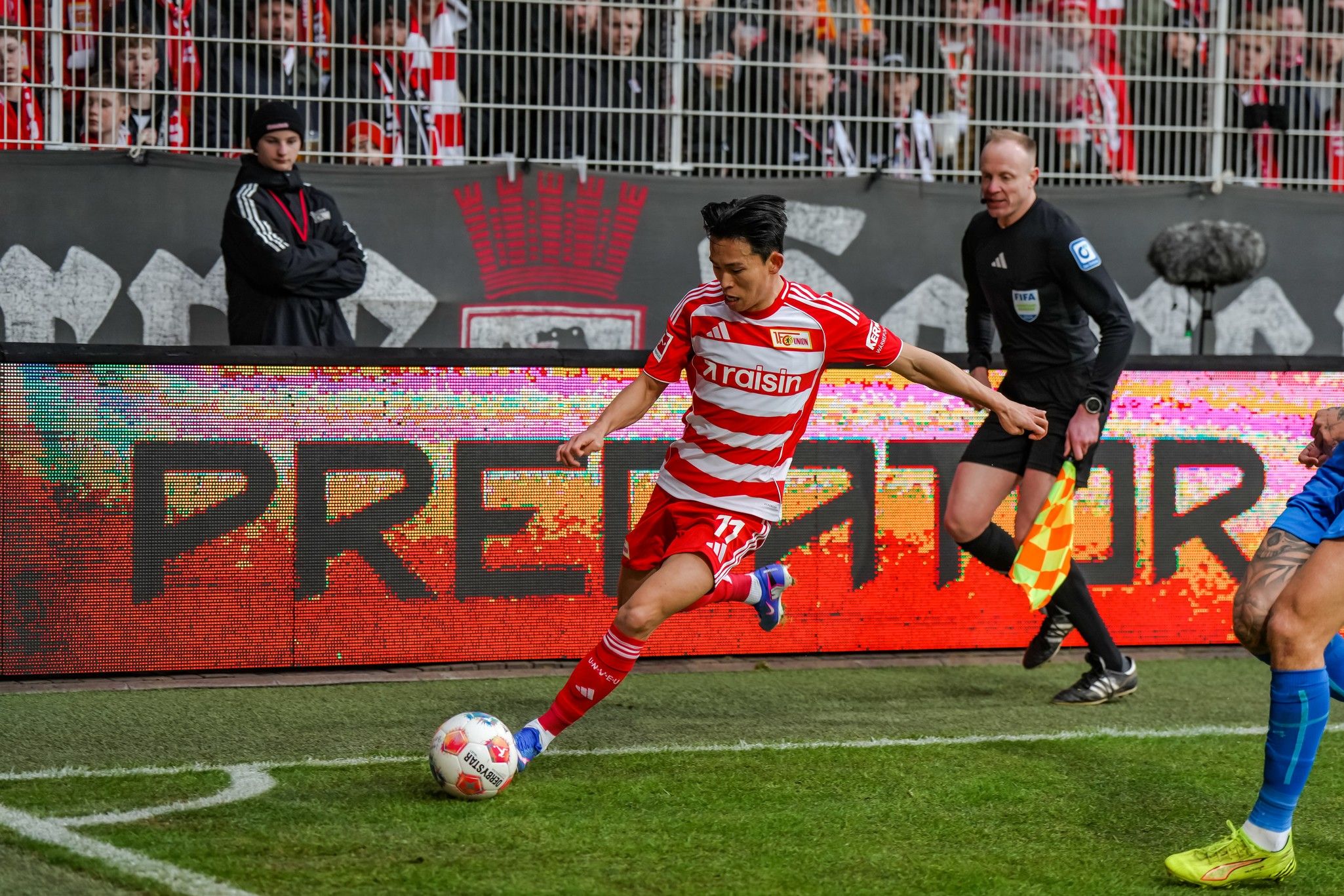 Spielszene: Ein Spieler in rot-weißer Kleidung führt einen Eckball aus, während der Schiedsrichter und andere Spieler beobachten.