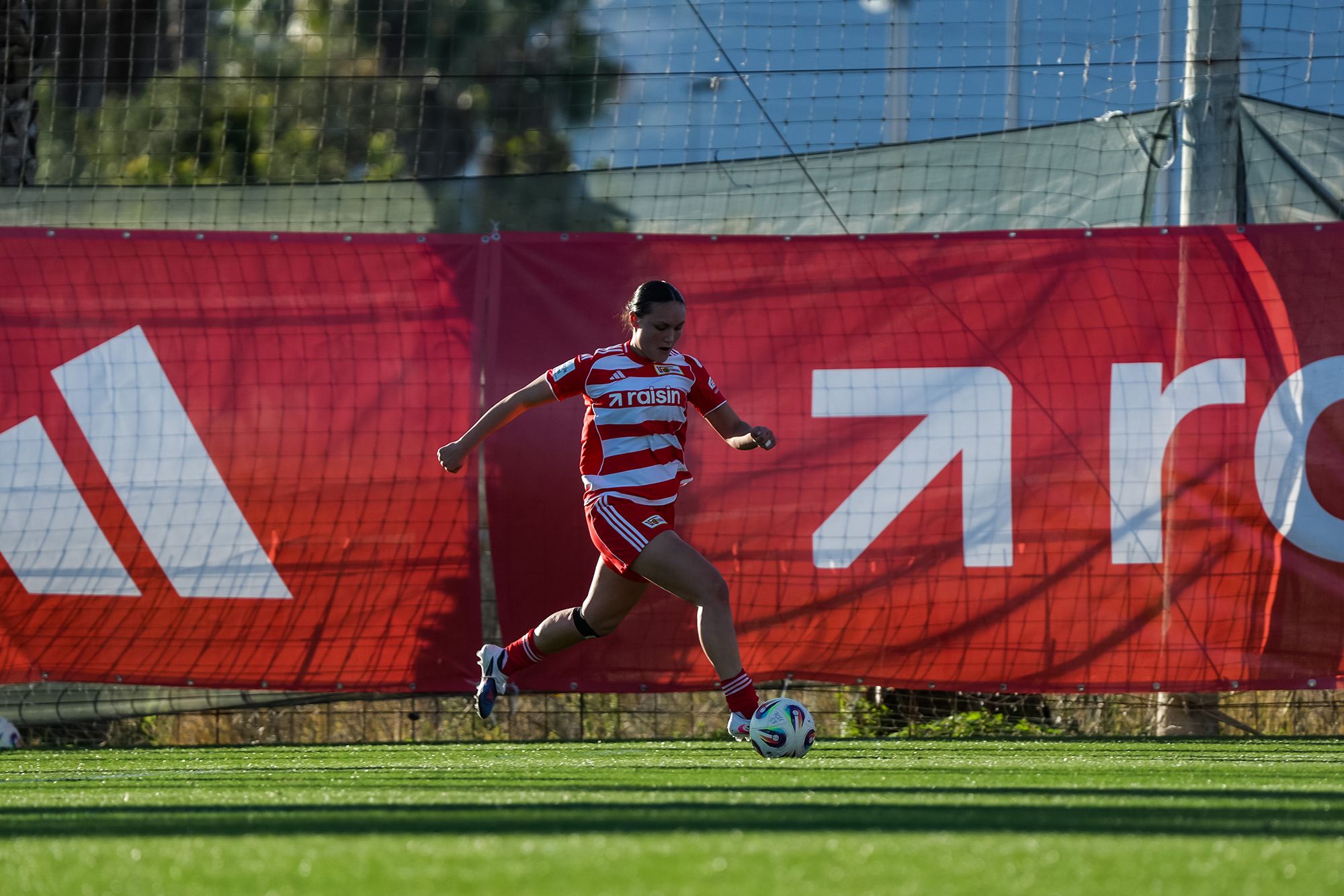Eine Spielerin in roten und weißen Streifen läuft auf dem Feld und spielt einen Ball, im Hintergrund ein Netz und eine Werbebande.