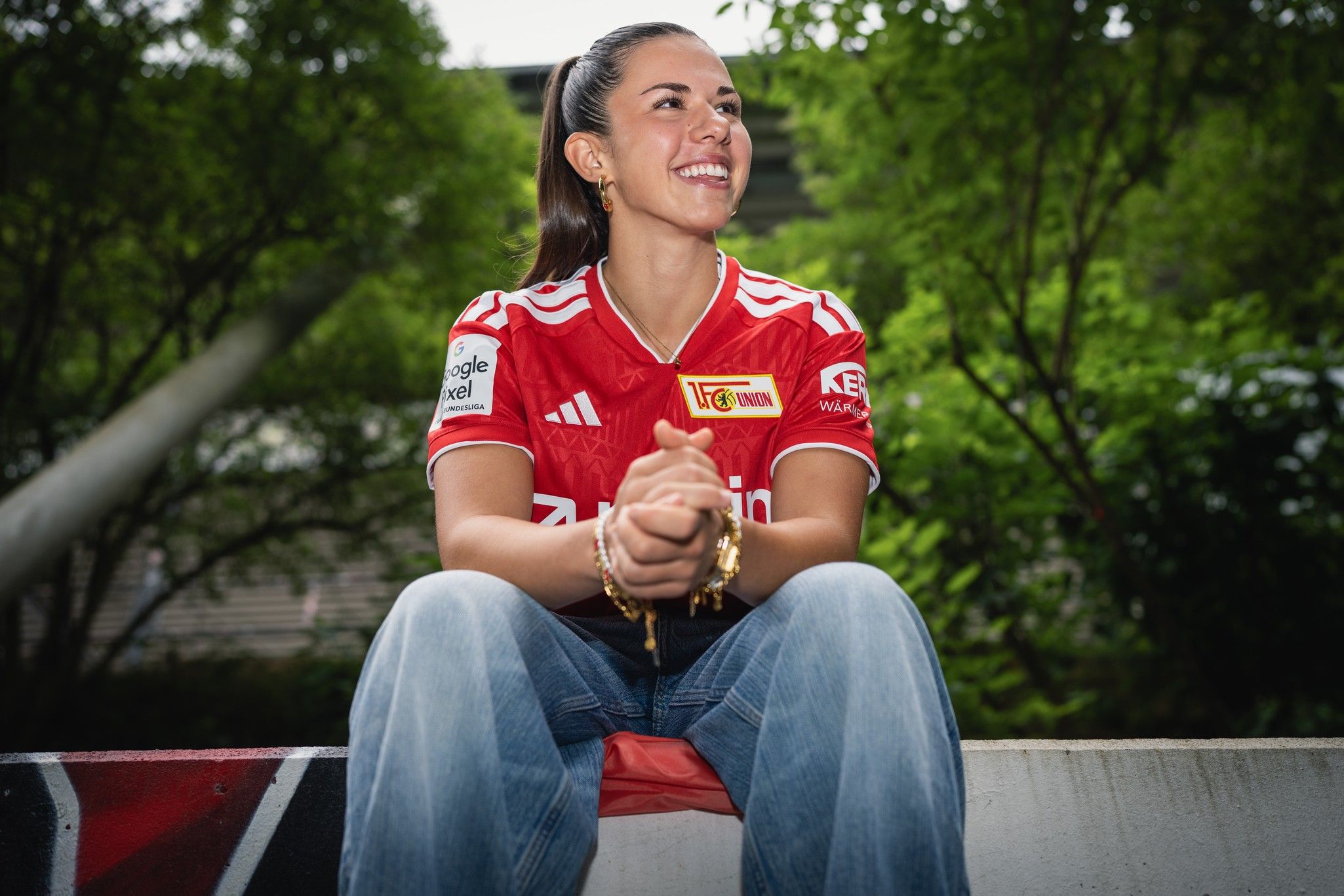 Eine lächelnde Person trägt ein rotes Fußballtrikot und sitzt entspannt auf einer Bank, umgeben von grüner Vegetation.
