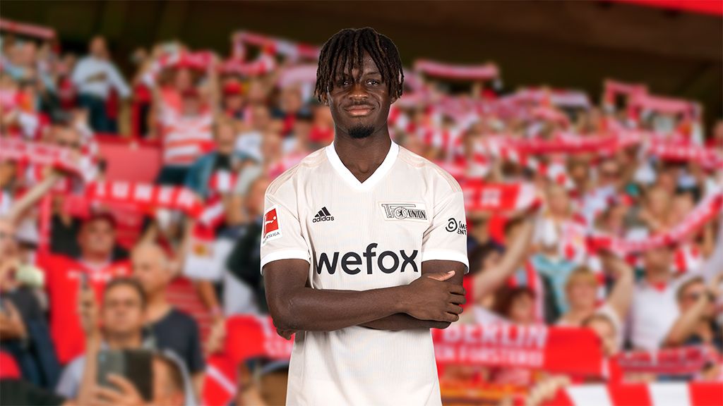 Ein Fußballspieler in einem weißen Trikot steht mit verschränkten Armen vor einer jubelnden Menge im Stadion.