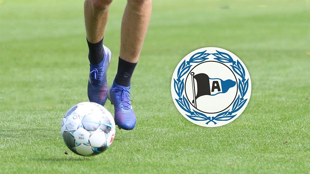 A player kicks a soccer ball on green grass, next to a club logo.