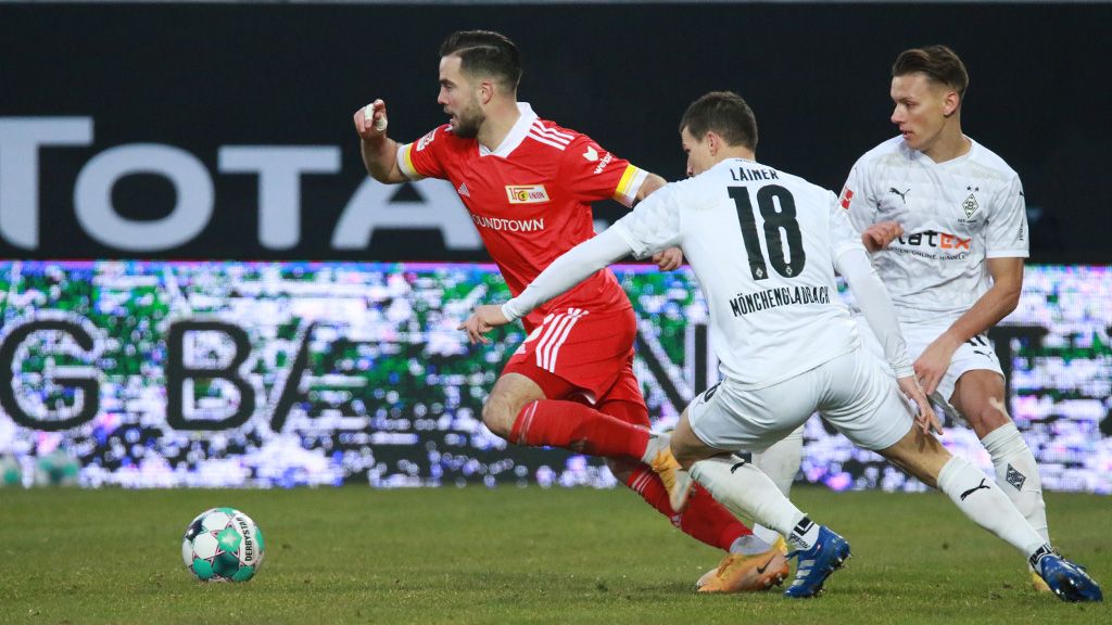 Ein Fußballspieler im roten Trikot dribbelt, während ein Gegner im weißen Trikot versucht, den Ball zu erobern.