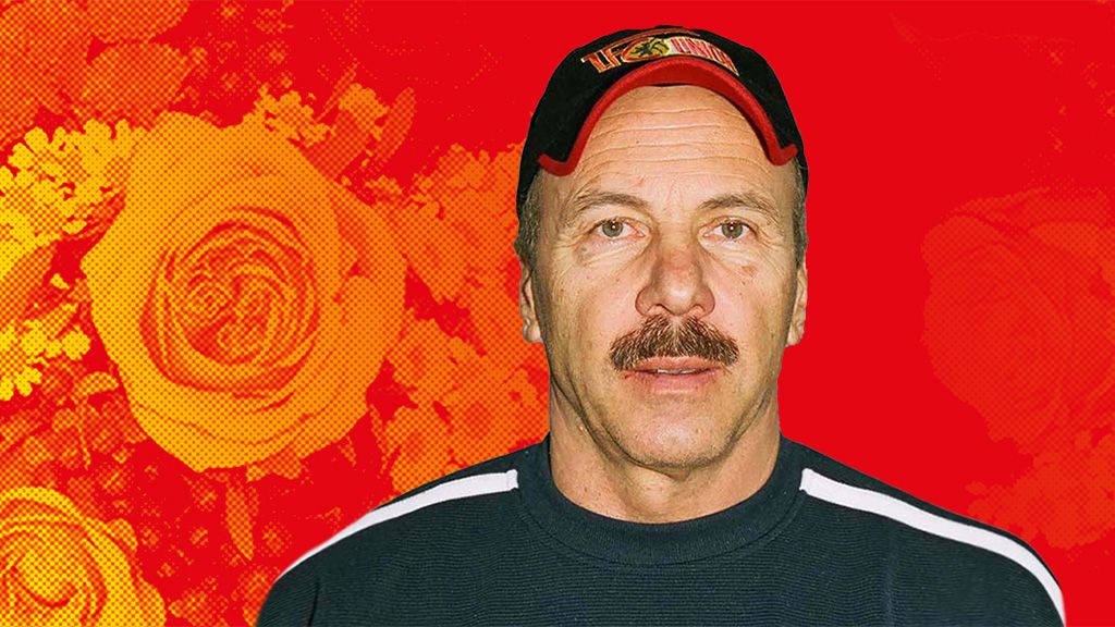 Ein Mann mit Schnurrbart trägt eine schwarze Mütze und ein dunkles Sweatshirt, vor einem rot-blumigen Hintergrund.