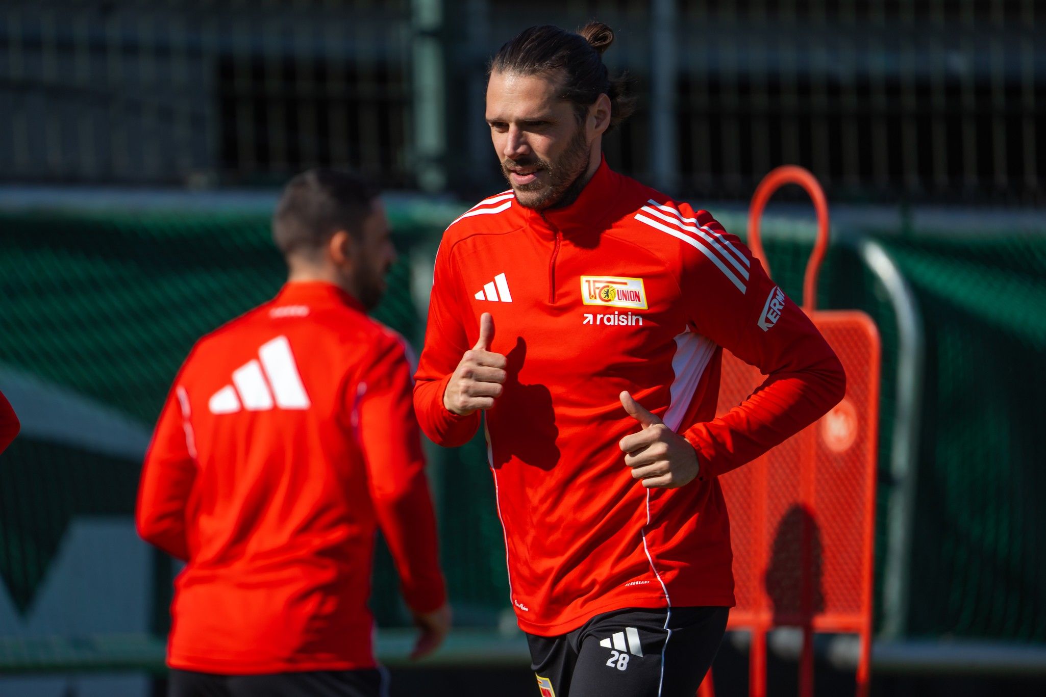 Ein Fußballspieler in rot-weißer Trainingskleidung joggt lächelnd und zeigt Daumen hoch. Andere Spieler im Hintergrund.