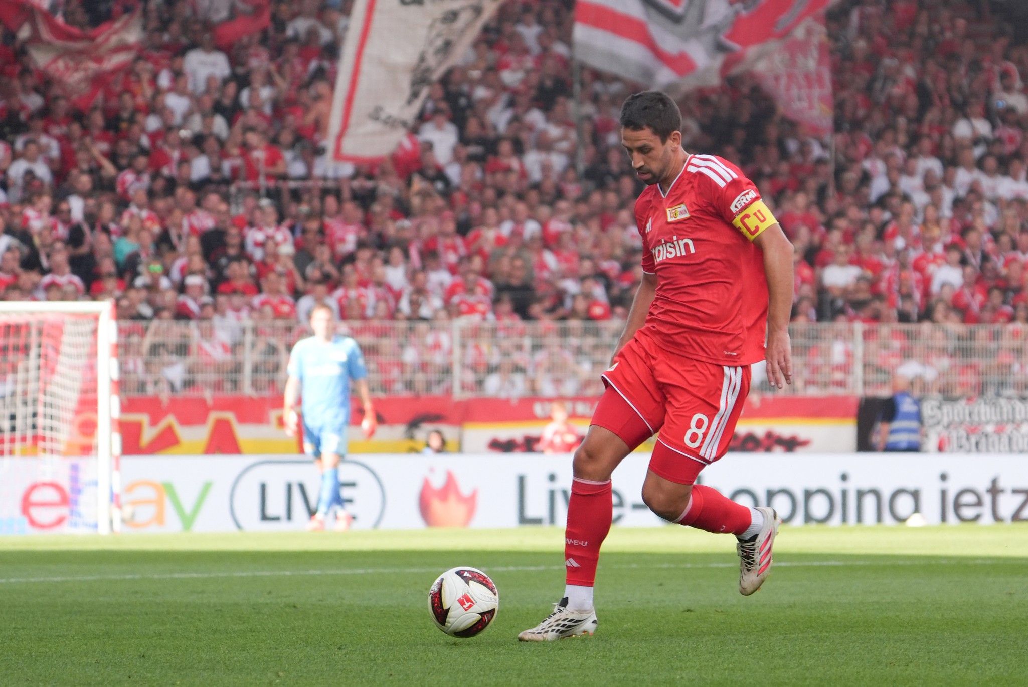Ein Fußballspieler in rotem Trikot mit der Nummer 8, der einen Ball in einem Stadion mit vielen Fans tritt.
