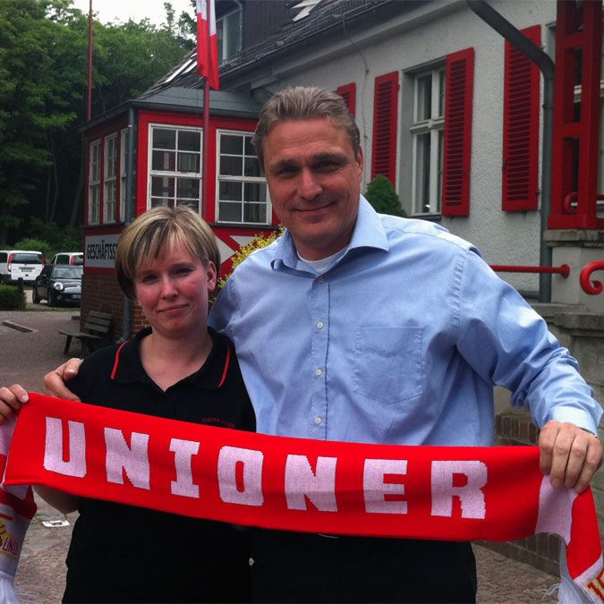 Zwei Personen stehen vor einem Gebäude und halten ein rotes Schal mit der Aufschrift "UNIONER".