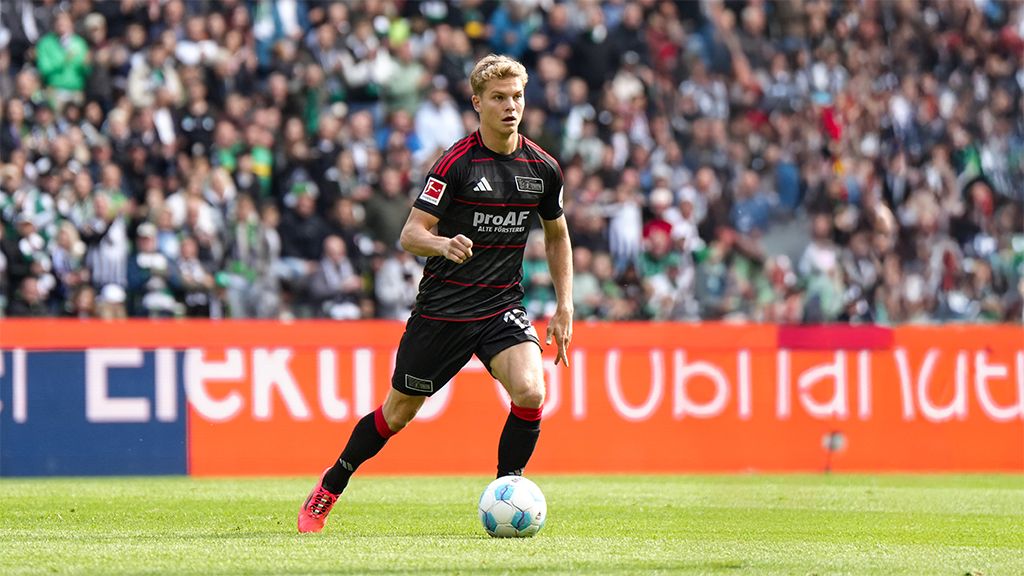 Fußballspieler in schwarzem Trikot dribbelt den Ball auf einem Stadionfeld, umgeben von jubelnden Fans.