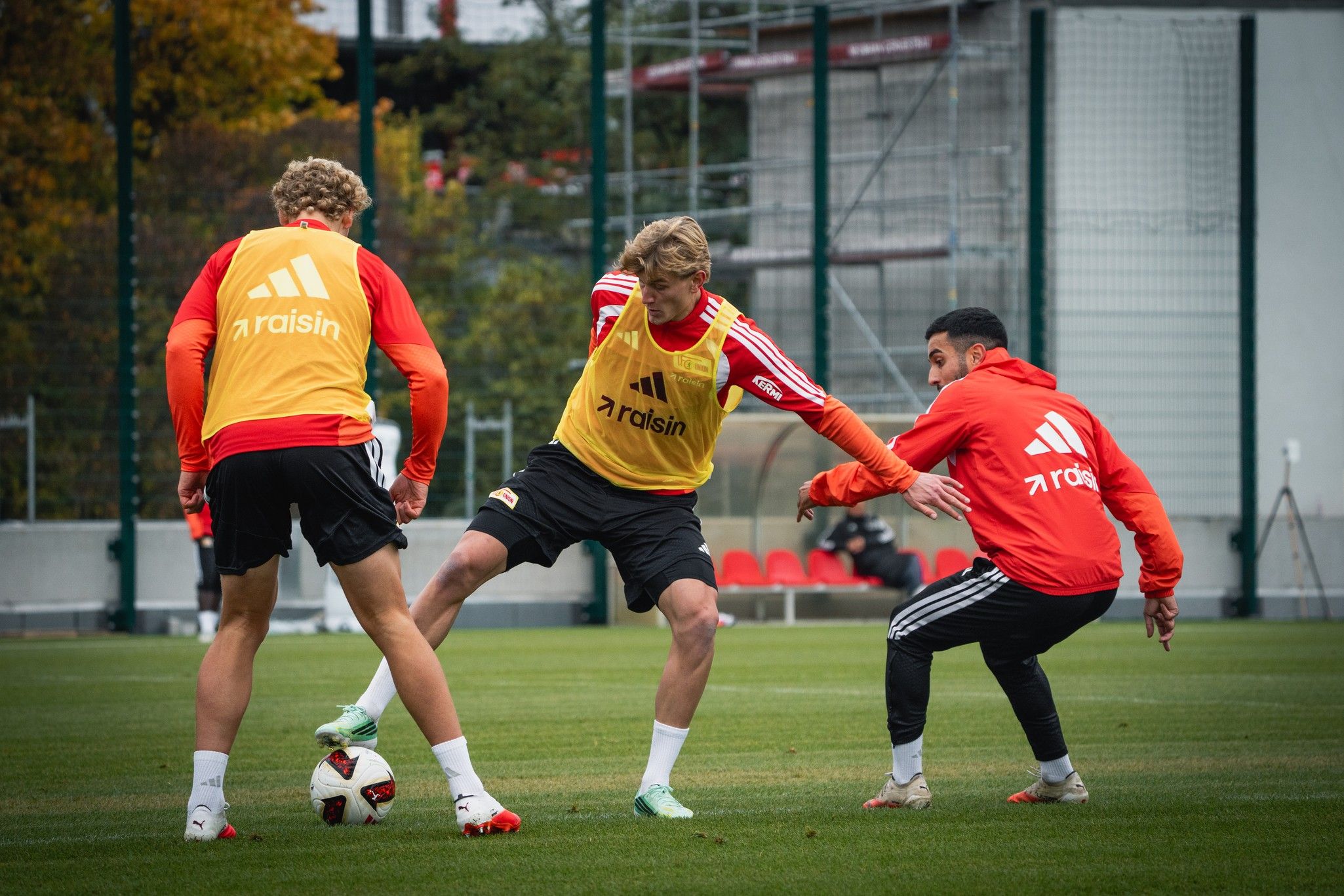 Drei Fußballspieler in Trainingskleidung üben auf einem Rasenplatz, während ein Spieler den Ball kontrolliert.