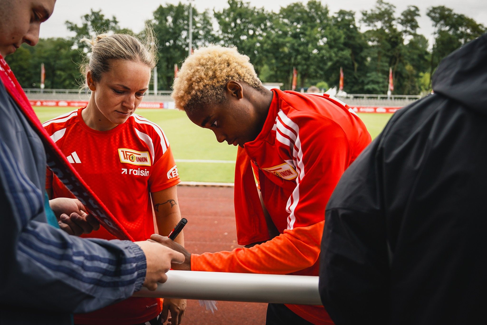 Zwei Sportler in roten Trikots unterzeichnen Autogramme auf einem Sportplatz.