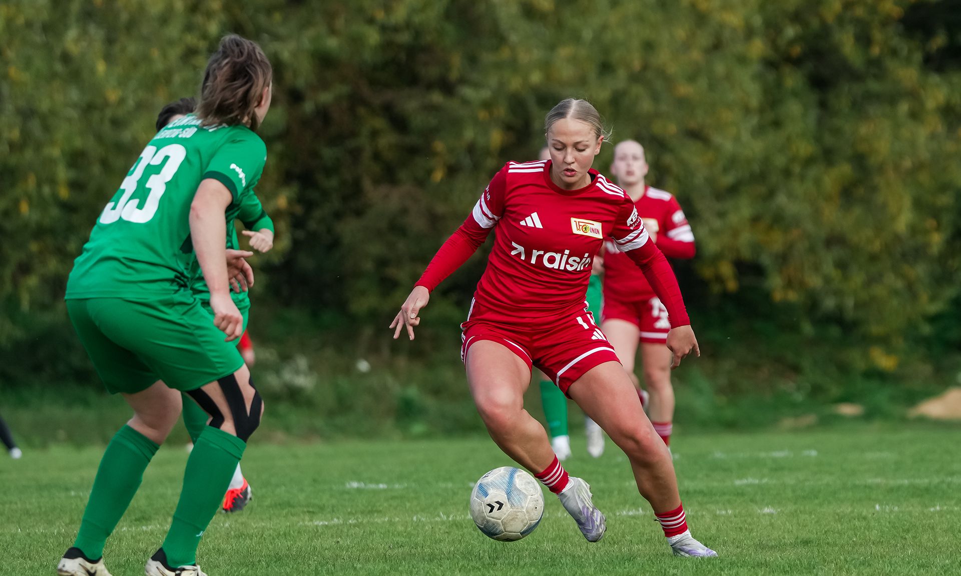 Eine Spielerin in rotem Trikot dribbelt mit einem Fußball auf einem grünen Rasenfeld, während eine Gegenspielerin in grün versucht, den Ball zu erreichen.