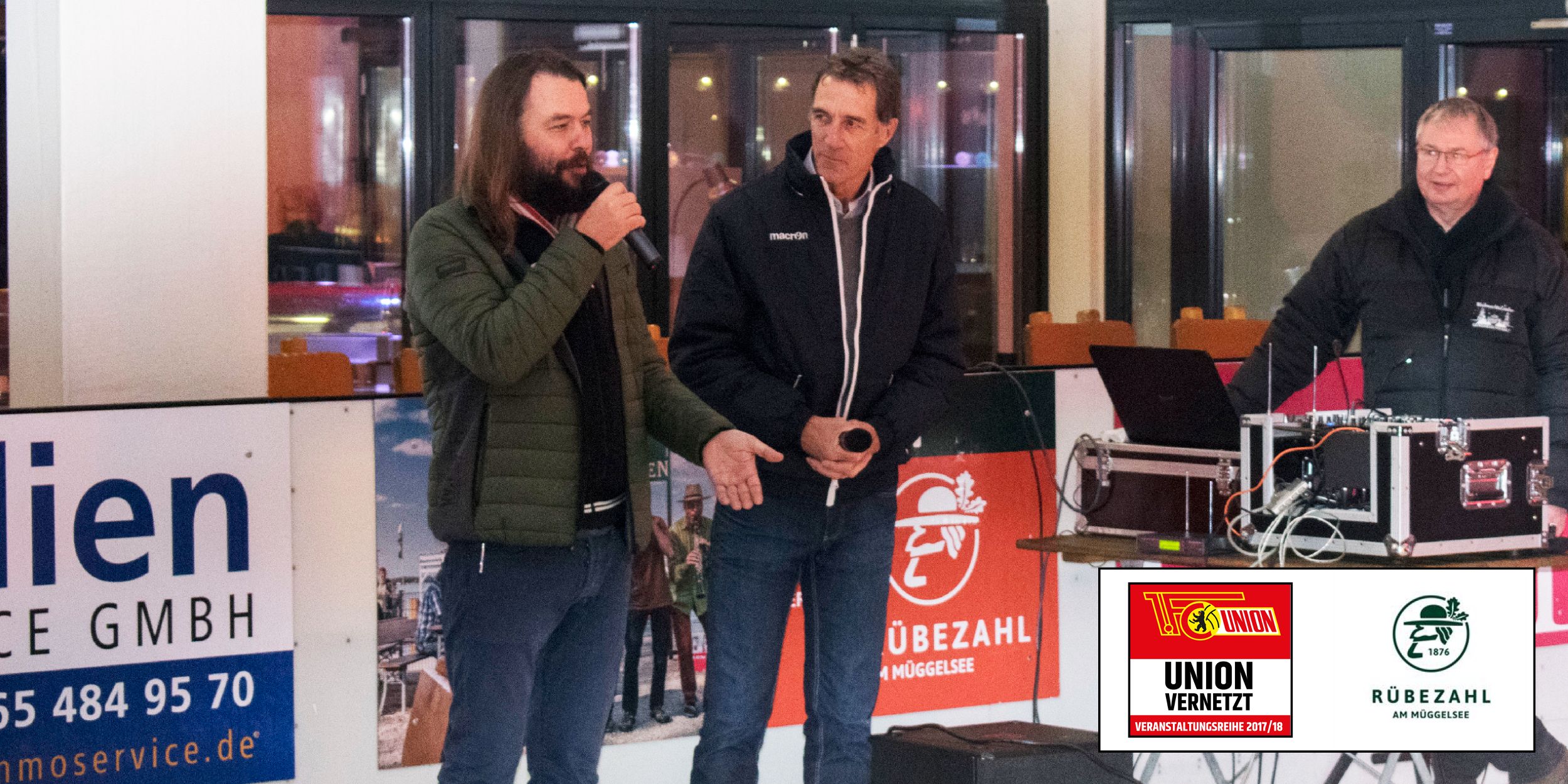 Zwei Männer sprechen auf einer Veranstaltung, während ein dritter Mann am Computer steht. Im Hintergrund Plakate und Banner.