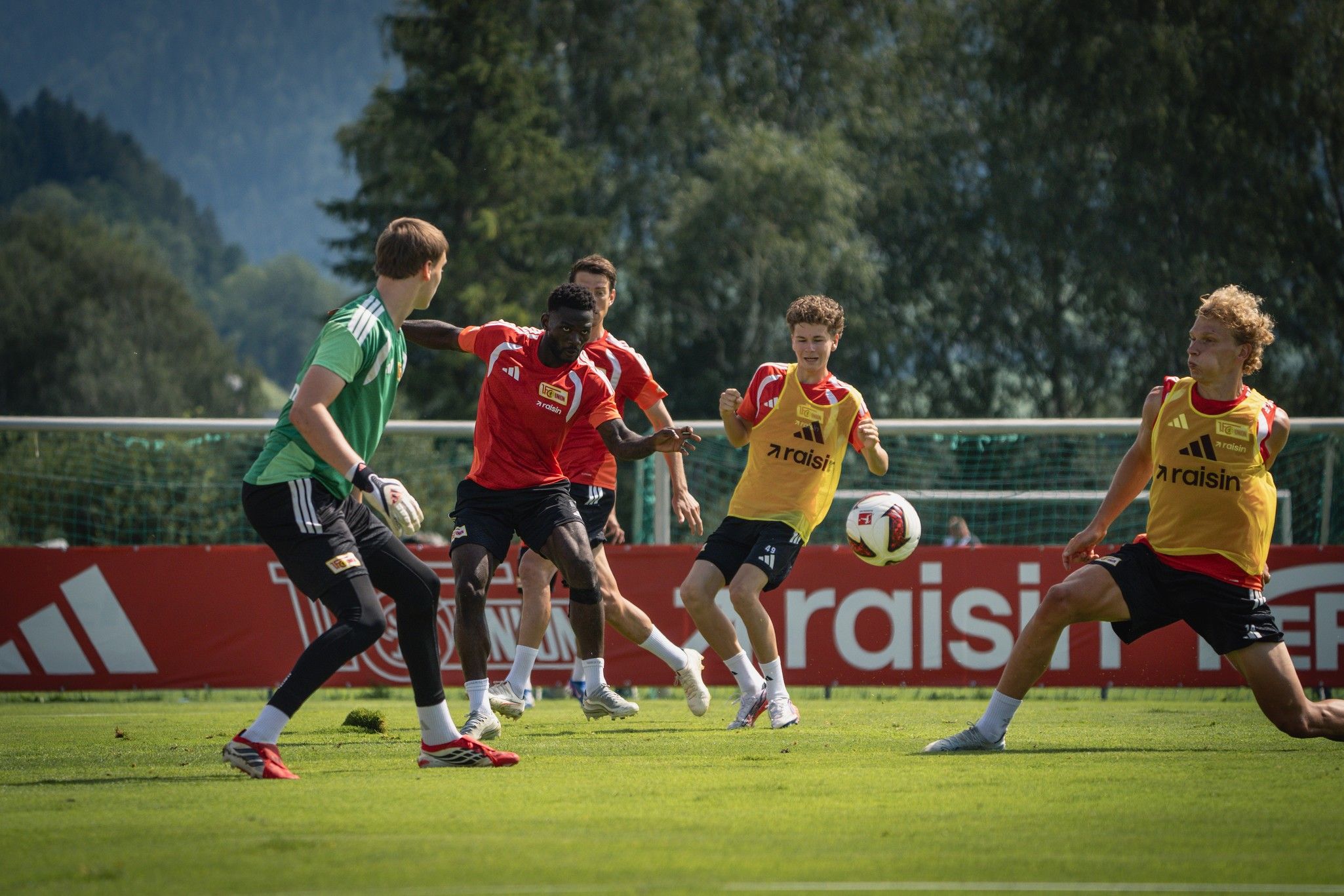 Fünf Fußballspieler in Trainingskleidung spielen auf einem grünen Feld, während ein Torwart im Hintergrund steht.