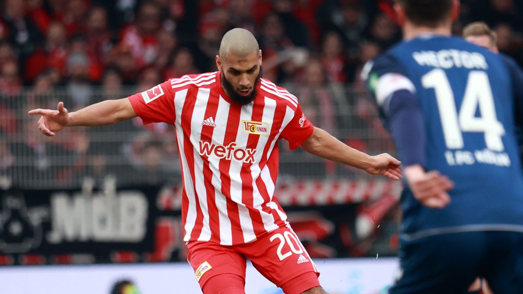 A football player in red and white club attire is in the game, while an opposing player in blue stands opposite him.