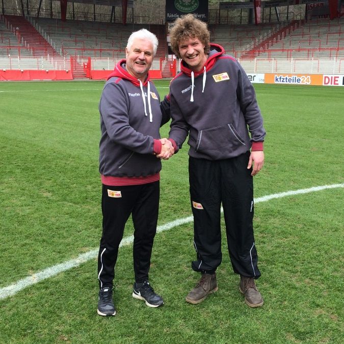 Zwei Männer in Sportkleidung schütteln sich die Hände auf einem Fußballfeld mit Rasen und Tribünen im Hintergrund.
