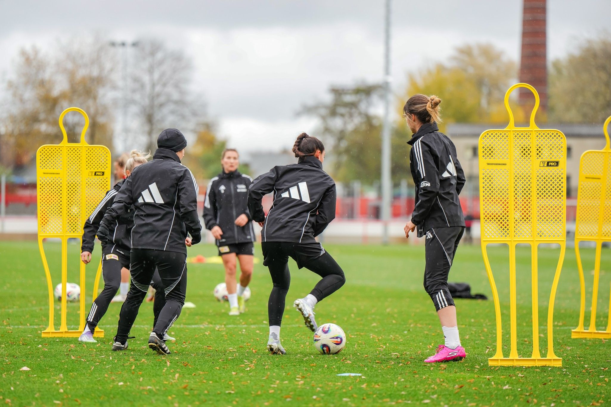 Frauenfußballmannschaft trainiert auf einem Platz mit gelben Trainingsdummies und einem Fußball bei bewölktem Wetter.