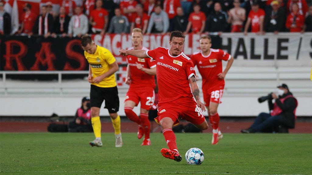 Ein Fußballspieler in rotem Trikot schießt auf das Tor, während Mitspieler und gegnerische Spieler im Hintergrund stehen.