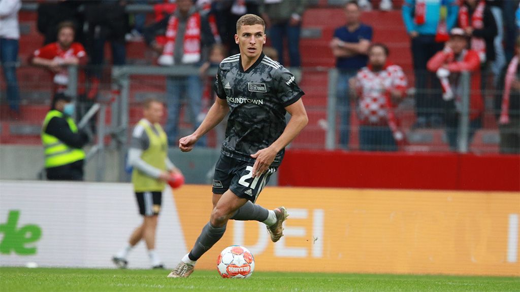 Spieler in grauem Fußballtrikot auf dem Spielfeld, mit Ball am Fuß, Zuschauer im Hintergrund.