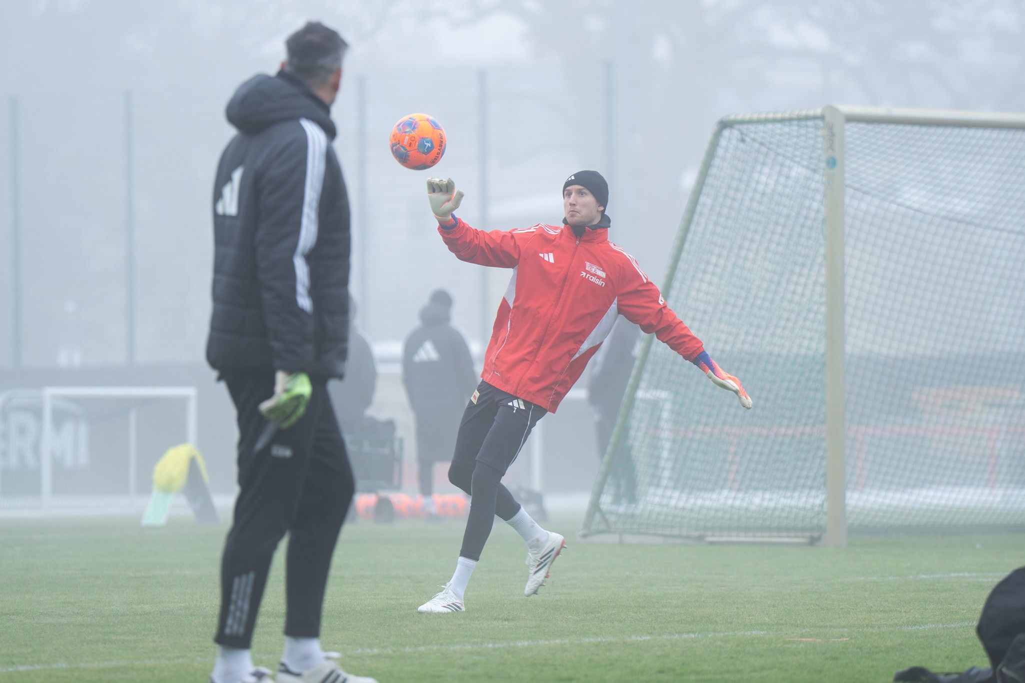 Torwart in roter Trainingsjacke fängt einen Ball während eines Trainings bei nebligem Wetter. Ein Trainer steht in der Nähe.