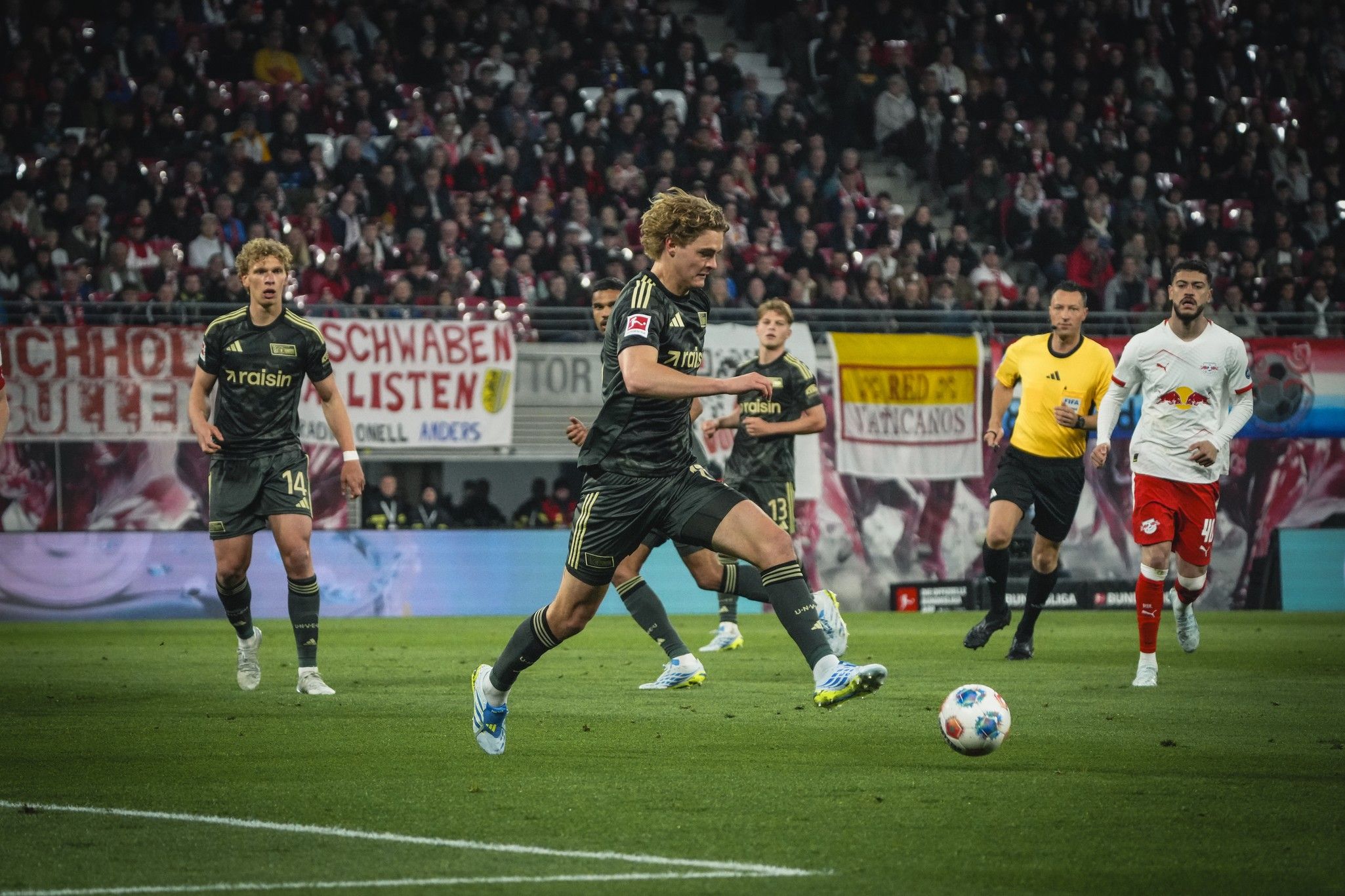 Fußballspieler in schwarzer Uniform schießt den Ball während eines Spiels, Zuschauer im Hintergrund.