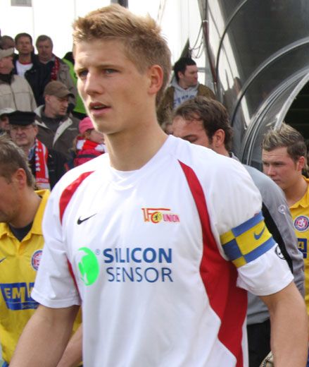 Ein Fußballspieler in einem weißen Trikot mit roten und gelben Akzenten, der Kapitänsbinde trägt, geht auf das Spielfeld.