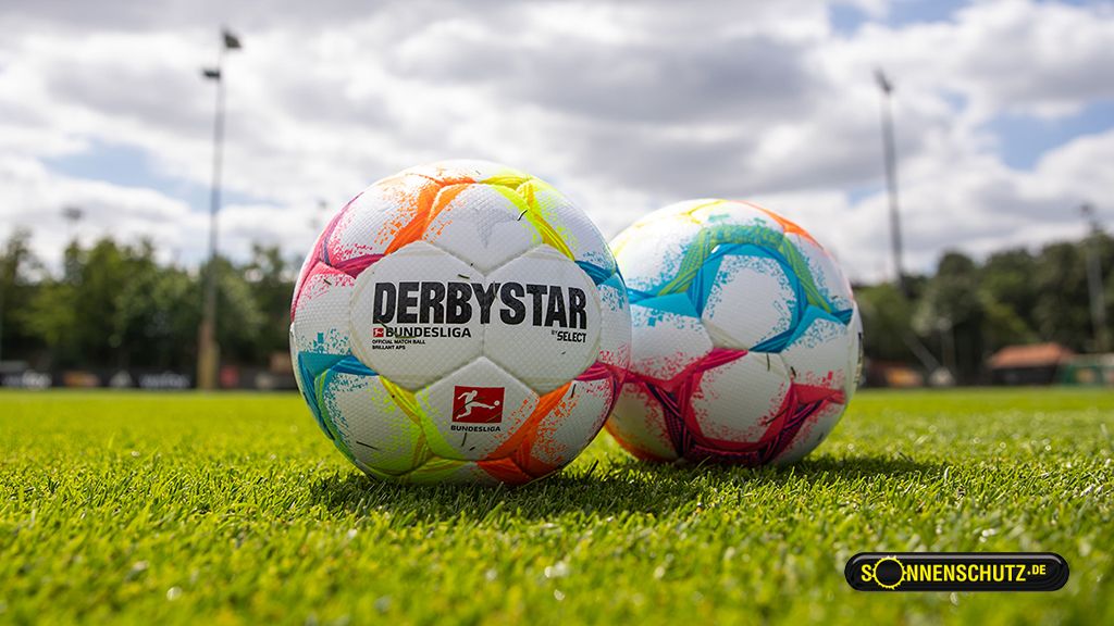 Zwei bunte Fußbälle liegen auf einem grünen Fußballfeld unter einem bewölkten Himmel.