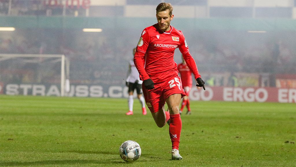Player in a red jersey dribbles the ball on the soccer field, with a blurred stadium atmosphere in the background.