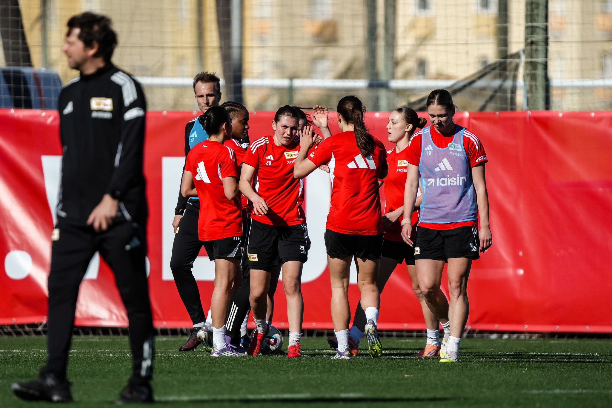 Eine Gruppe von Sportlerinnen in roten Trikots trainiert auf einem Fußballfeld, während ein Trainer im Hintergrund steht.