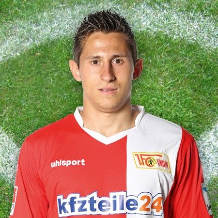 Ein junger Fußballspieler in einem roten und weißen Trikot steht vor einem grünen Rasenhintergrund.