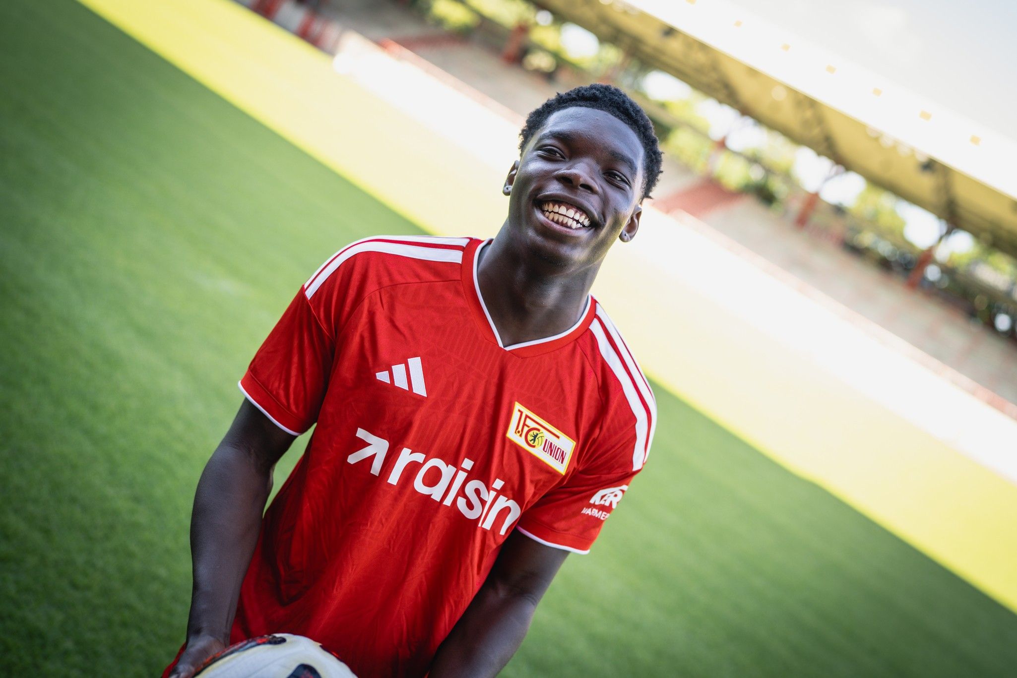 Ein junger Fußballspieler in rotem Trikot lächelt und hält einen Ball in einem Stadion.