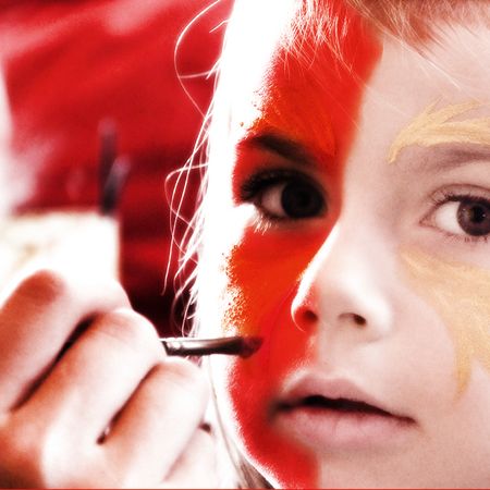 Ein Kind mit halbem roten und halbem goldenen Gesicht, während es von jemandem bemalt wird. Künstliche Hand mit Pinsel sichtbar.