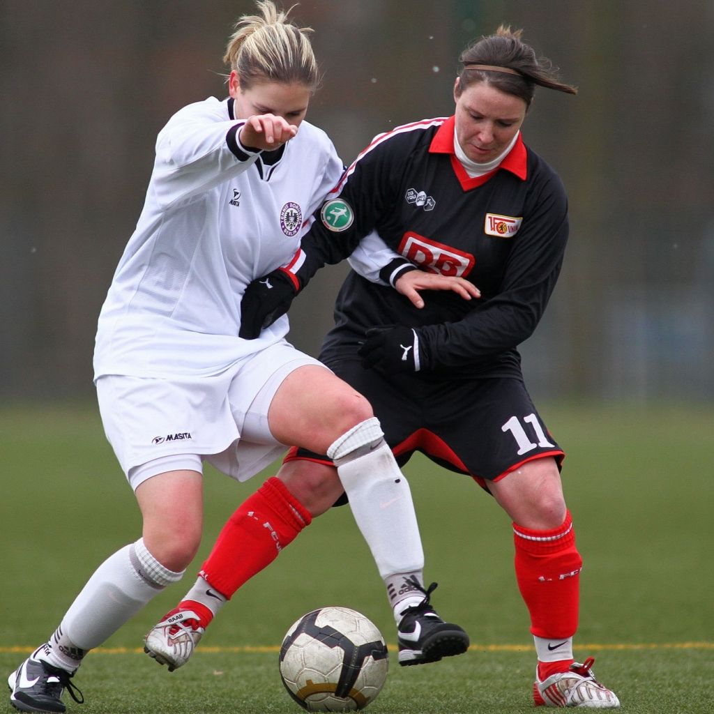 Zwei Frauen im Fußballspiel, eine in weißem Trikot mit Schiedsrichterarmbinde, die andere in schwarzem Trikot, kämpfen um den Ball.