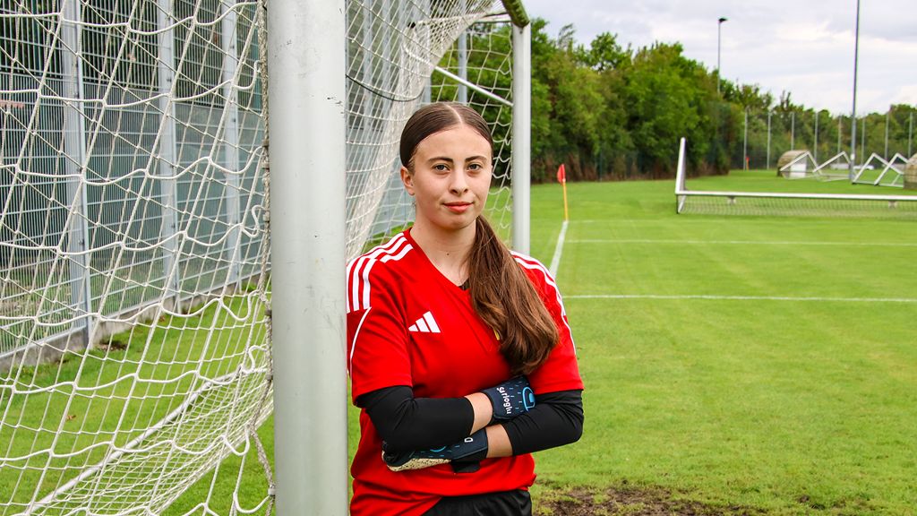 Junge Torhüterin in rotem Trikot steht lächelnd neben einem Fußballtor auf einem Rasenplatz.