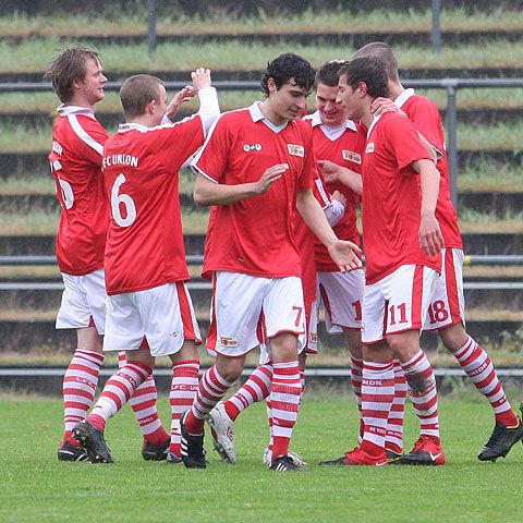 Sechs Fußballspieler in roten Trikots feiern einen Torerfolg auf dem Platz.