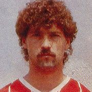 Ein Mann mit lockigem, braunem Haar und Bart, trägt ein rotes Sporttrikot. Hintergrund ist hell und neutral.