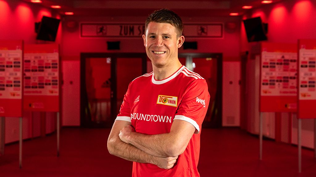 Ein lächelnder Fußballspieler in rotem Trikot mit dem Aufdruck "1. FC Union" steht vor einem roten Hintergrund.