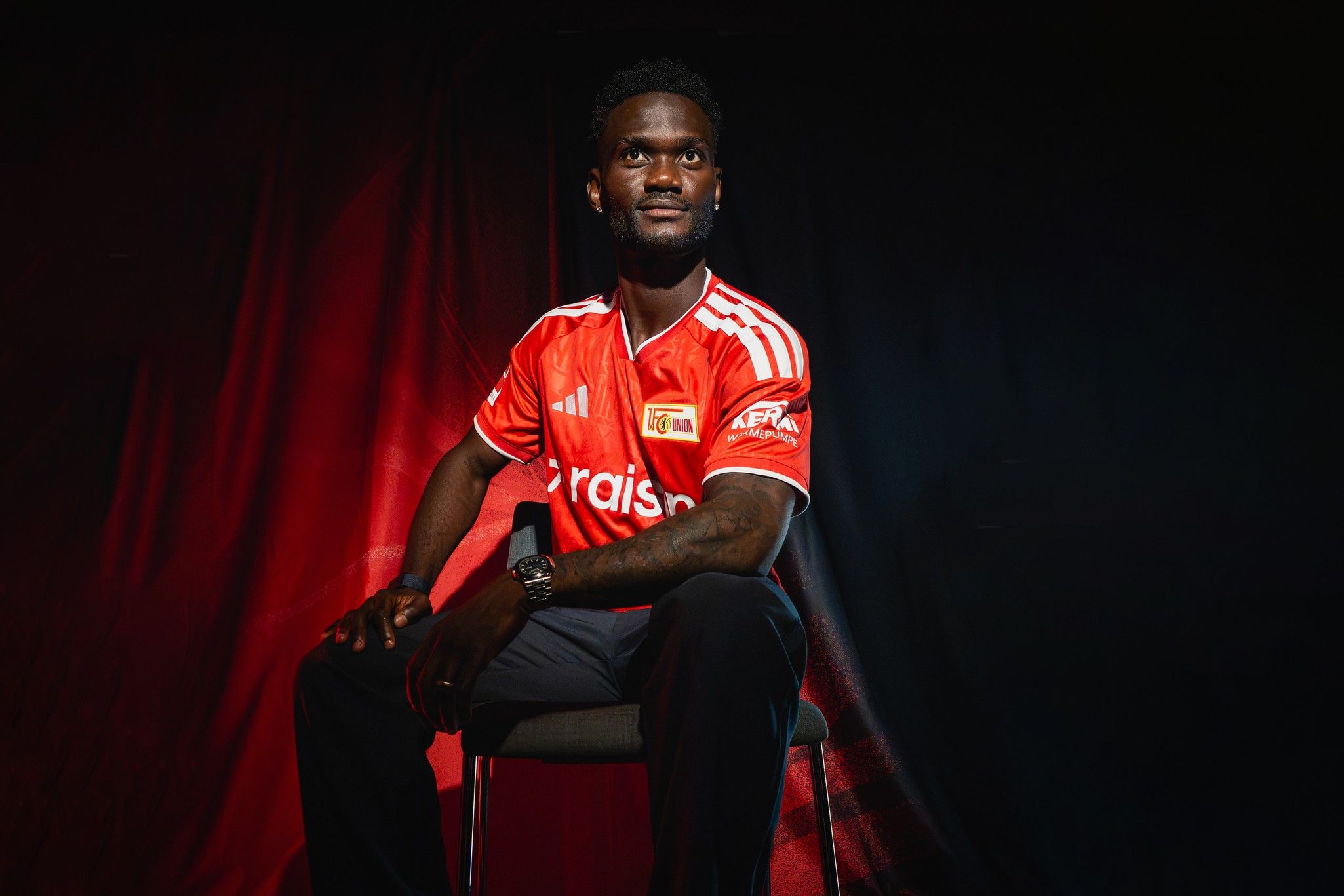 Ein Mann mit lächelndem Gesicht trägt ein rotes Fußballtrikot und sitzt auf einem Stuhl vor einem dunklen Hintergrund.