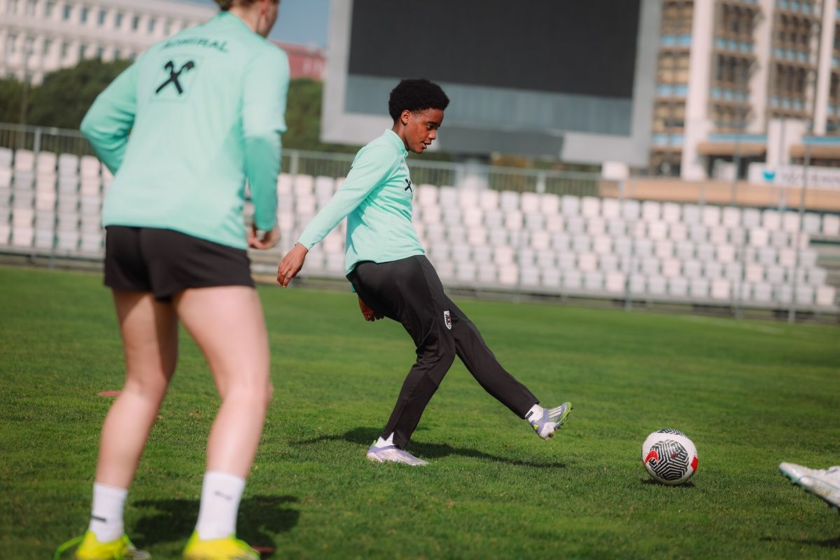 Zwei Fußballspieler trainieren auf einem Rasenplatz, einer tritt einen Ball. Beide tragen sportliche Oberteile und Shorts.