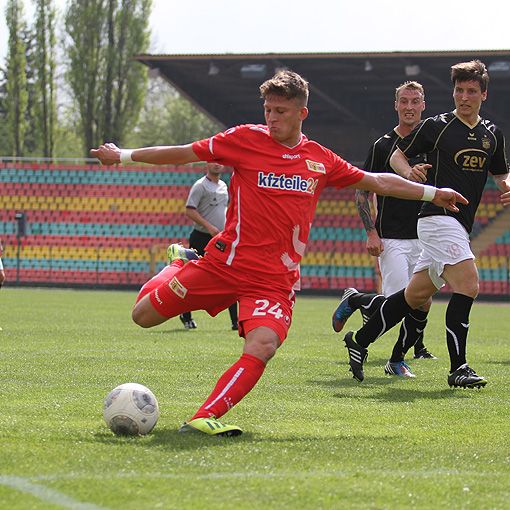 Ein Spieler in rotem Trikot schießt einen Fußball, während zwei gegnerische Spieler in schwarz-wei�ßen Trikots im Hintergrund stehen.