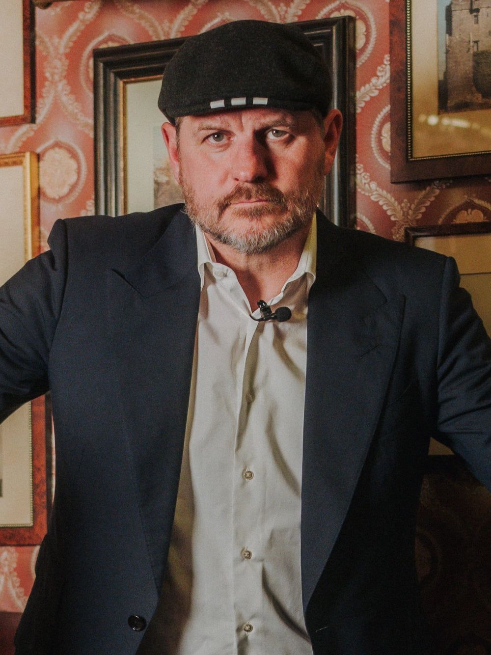 Mann mit Bart und Kappe in einem Anzug vor einer mit Bildern dekorierten Wand.