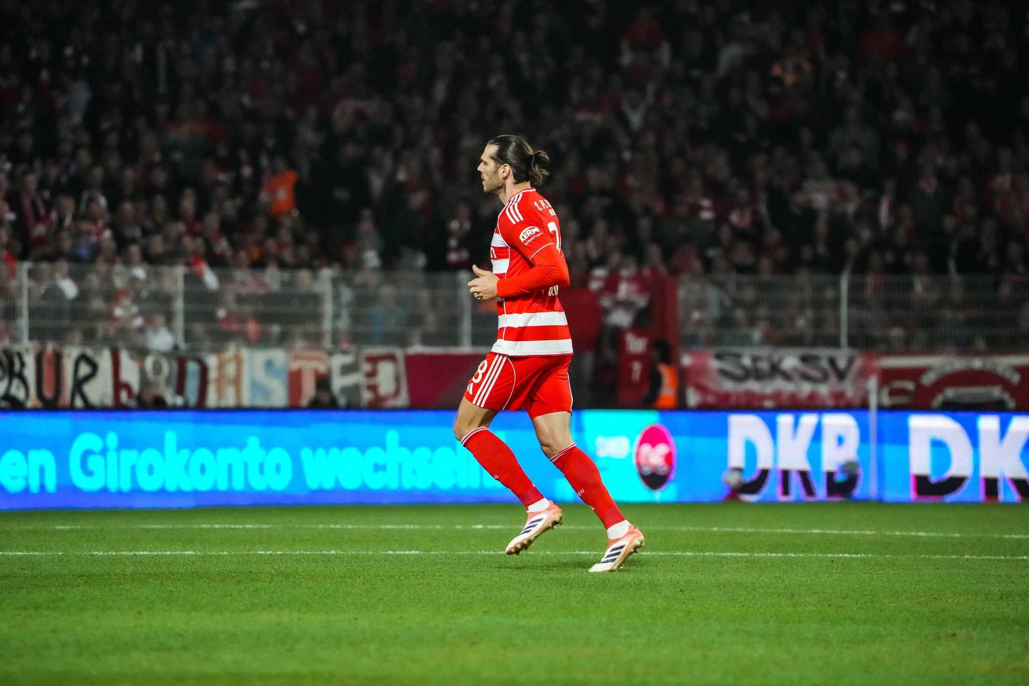 Ein Fußballspieler in rotem Trikot sprintet auf dem Spielfeld, umgeben von jubelnden Fans im Stadion.