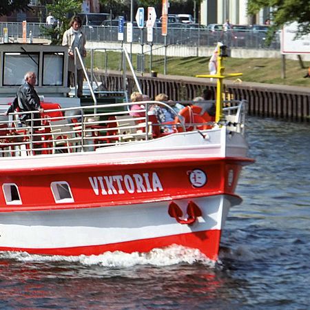 Ein rotes Ausflugsschiff namens "Viktoria" fährt auf einem Gewässer, Passagiere sitzen an Deck und genießen die Fahrt.