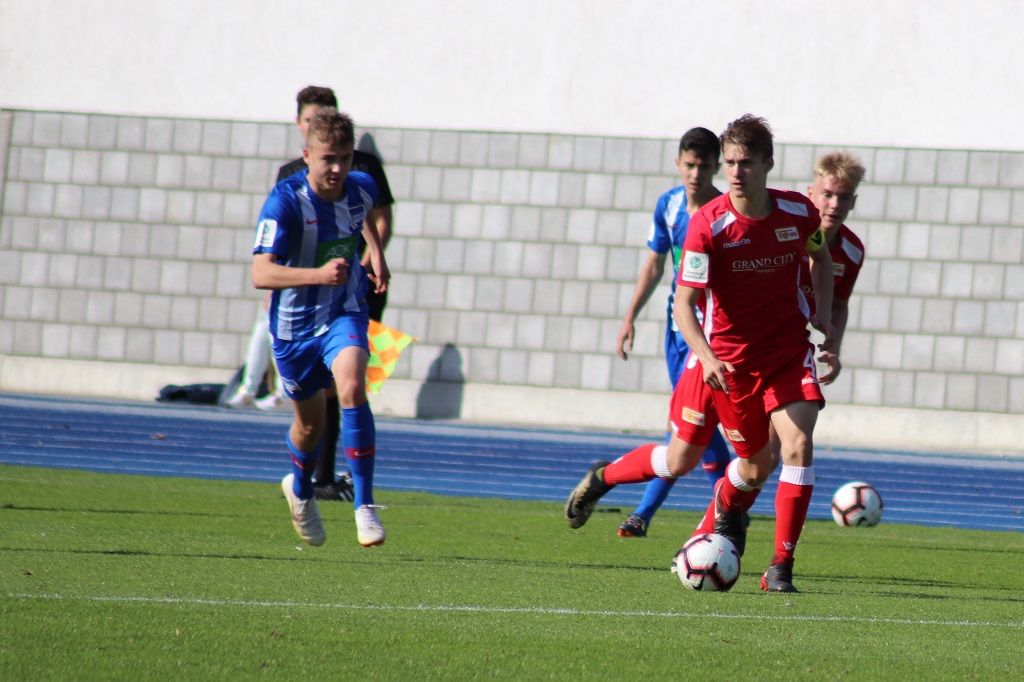 Fußballspiel mit Spielern in roten und blauen Trikots auf einem Rasenplatz. Ein Spieler dribbelt den Ball.
