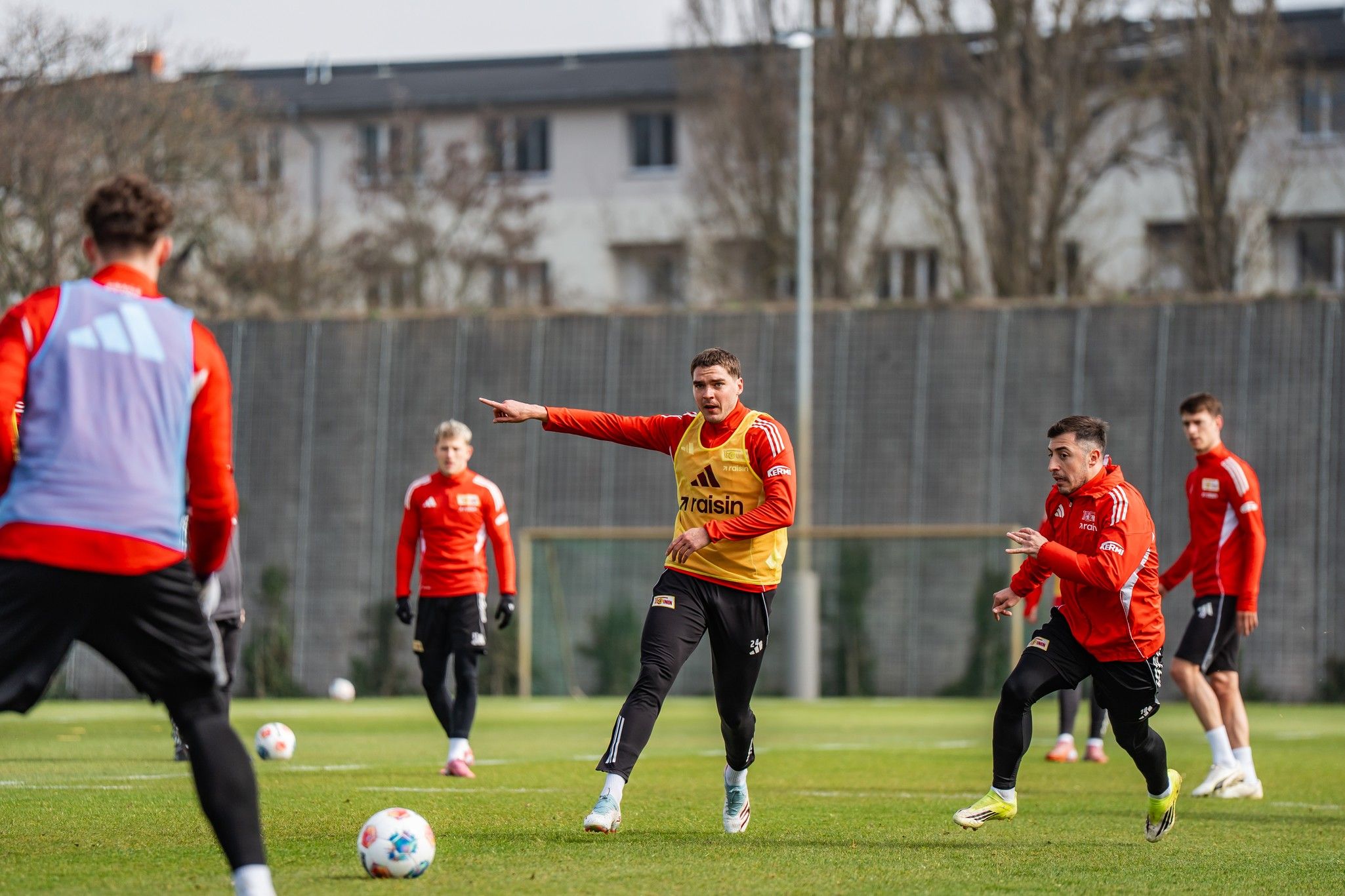 Spieler in Sportkleidung trainieren auf einem Fußballplatz, während ein Spieler Anweisungen gibt. Hintergrund mit Bäumen und Gebäuden.